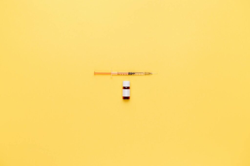 Minimalist image of a syringe and vaccine vial on a vibrant yellow background.